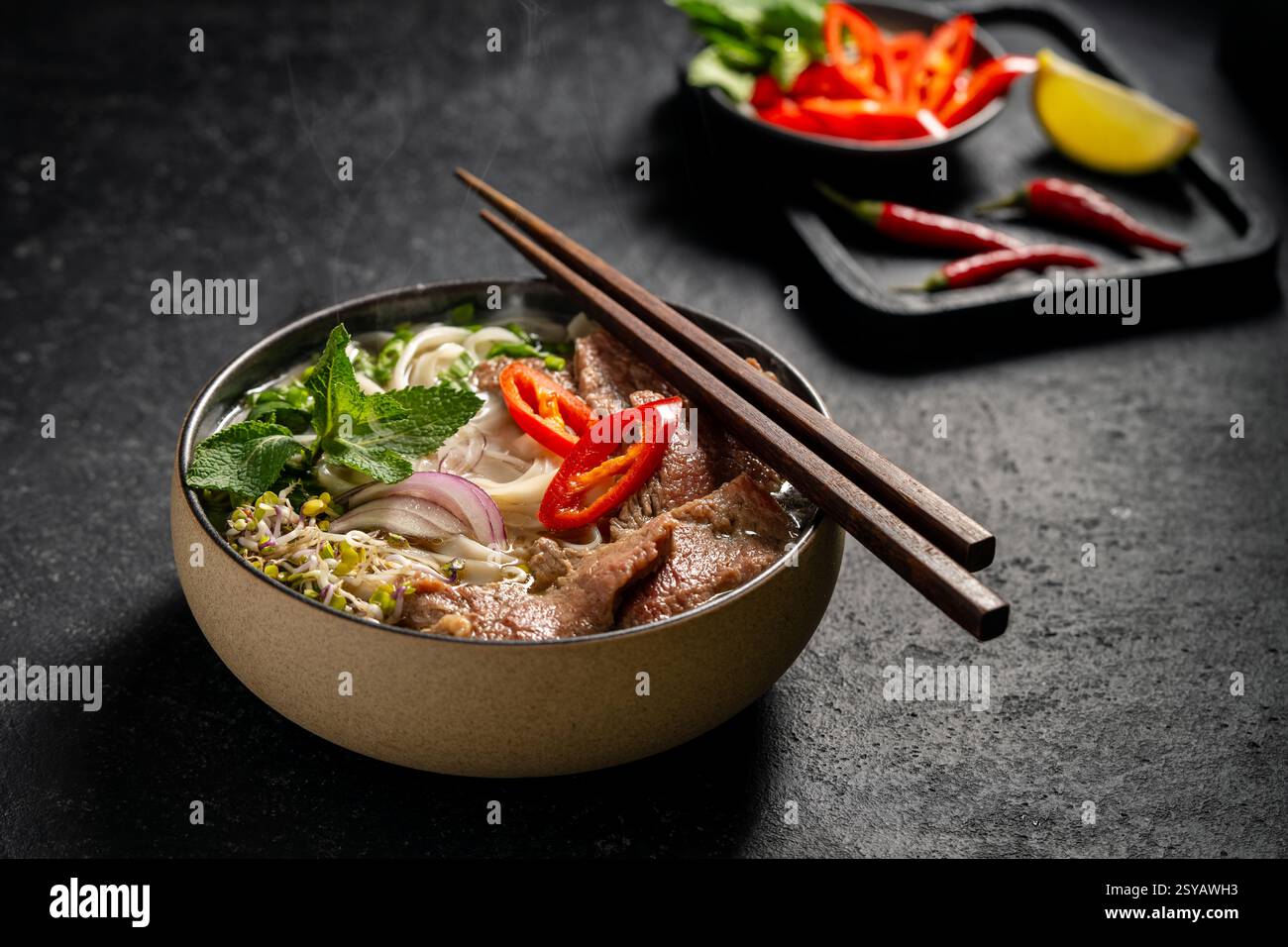 Traditionelle vietnamesische Rindfleisch-Pho-Nudelsuppe in einer Keramikschale, garniert mit Rindfleischscheiben, Kräutern, roter Chili und Limette auf dunklem Steinhintergrund. Stockfoto