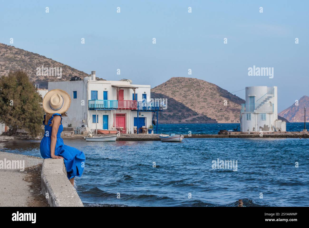 Ein ruhiges Küstendorf in Empourios auf der Insel Milos, Griechenland. Eine Frau in blauem Kleid bewundert die Aussicht mit traditionellen weißen Gebäuden und Booten Stockfoto