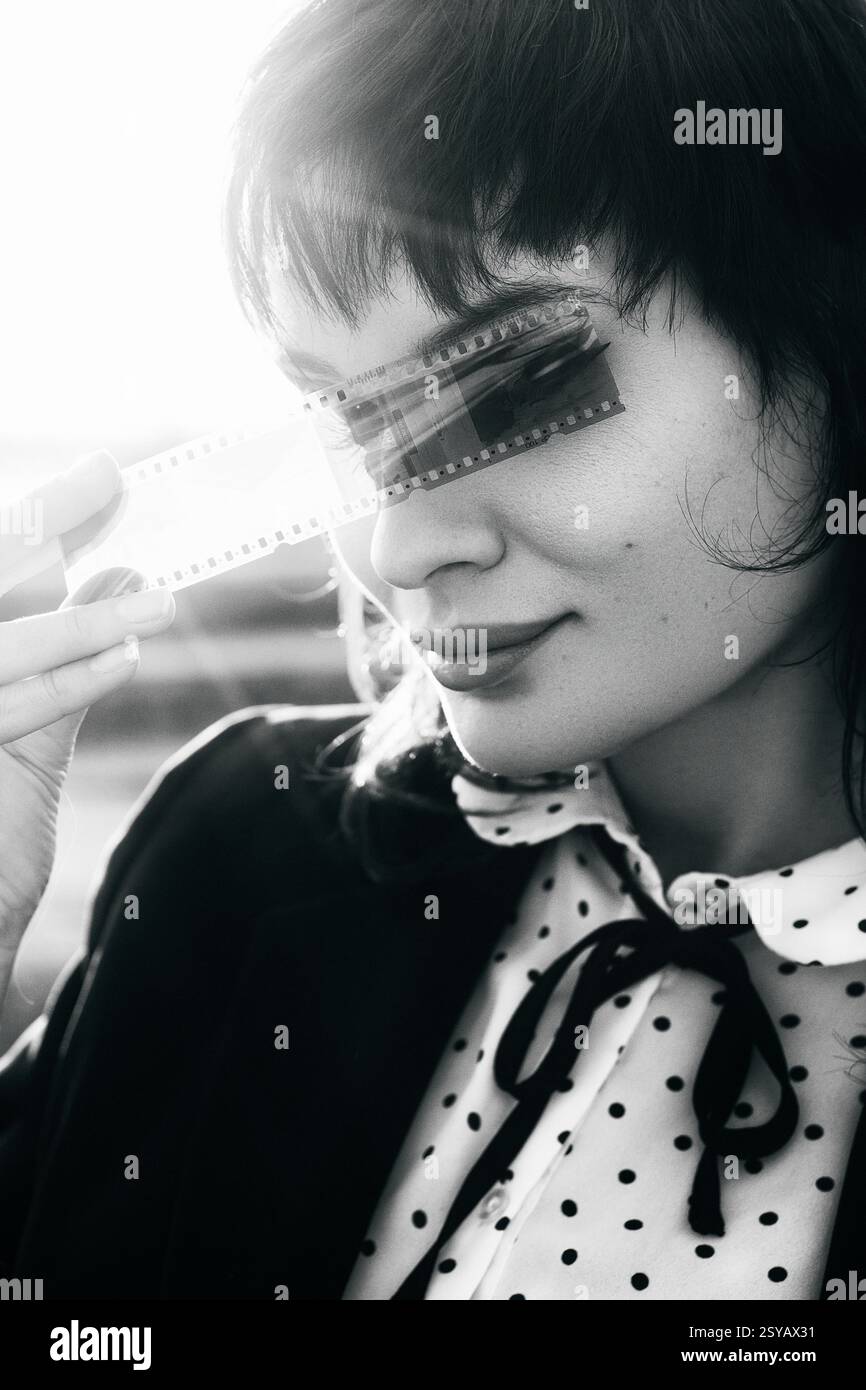 Schwarz-weiß-Porträt einer Frau, die einen Filmstreifen über den Augen hält, was ein Gefühl von Mysterium und Nostalgie weckt, mit Sonnenlicht im Hintergrund. Stockfoto