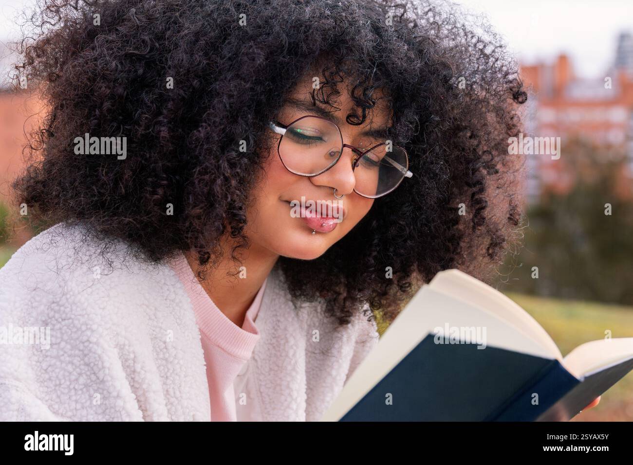 Eine junge lateinfrau mit lockigem Haar und Brille liest draußen ein Buch und genießt eine friedliche und entspannende Umgebung. Sie ist lässig gekleidet und Appa Stockfoto