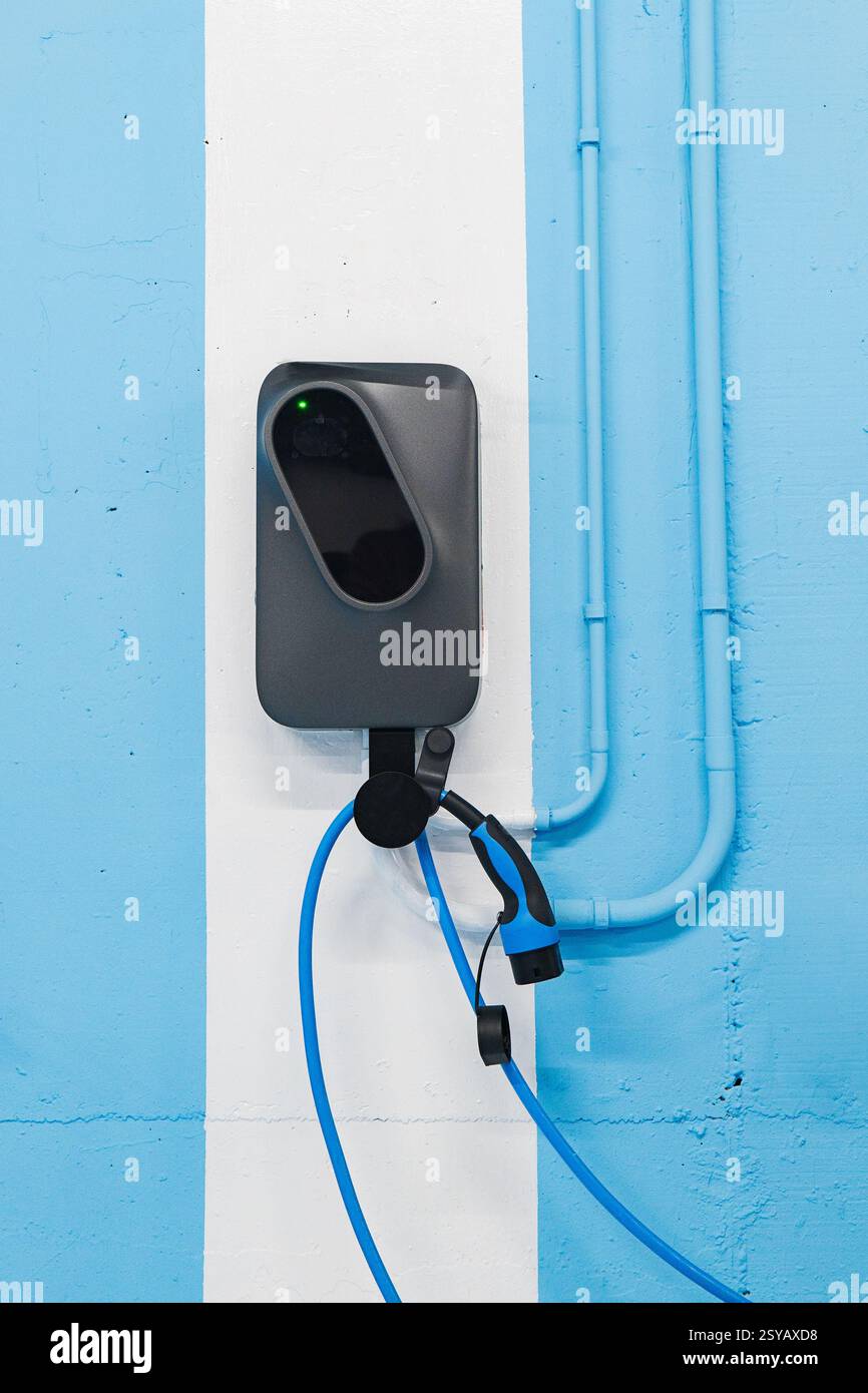 Eine Ladestation für Elektrofahrzeuge, die an einer hellblau-weiß gestreiften Wand mit sichtbaren Rohren und Drähten montiert ist. Stockfoto