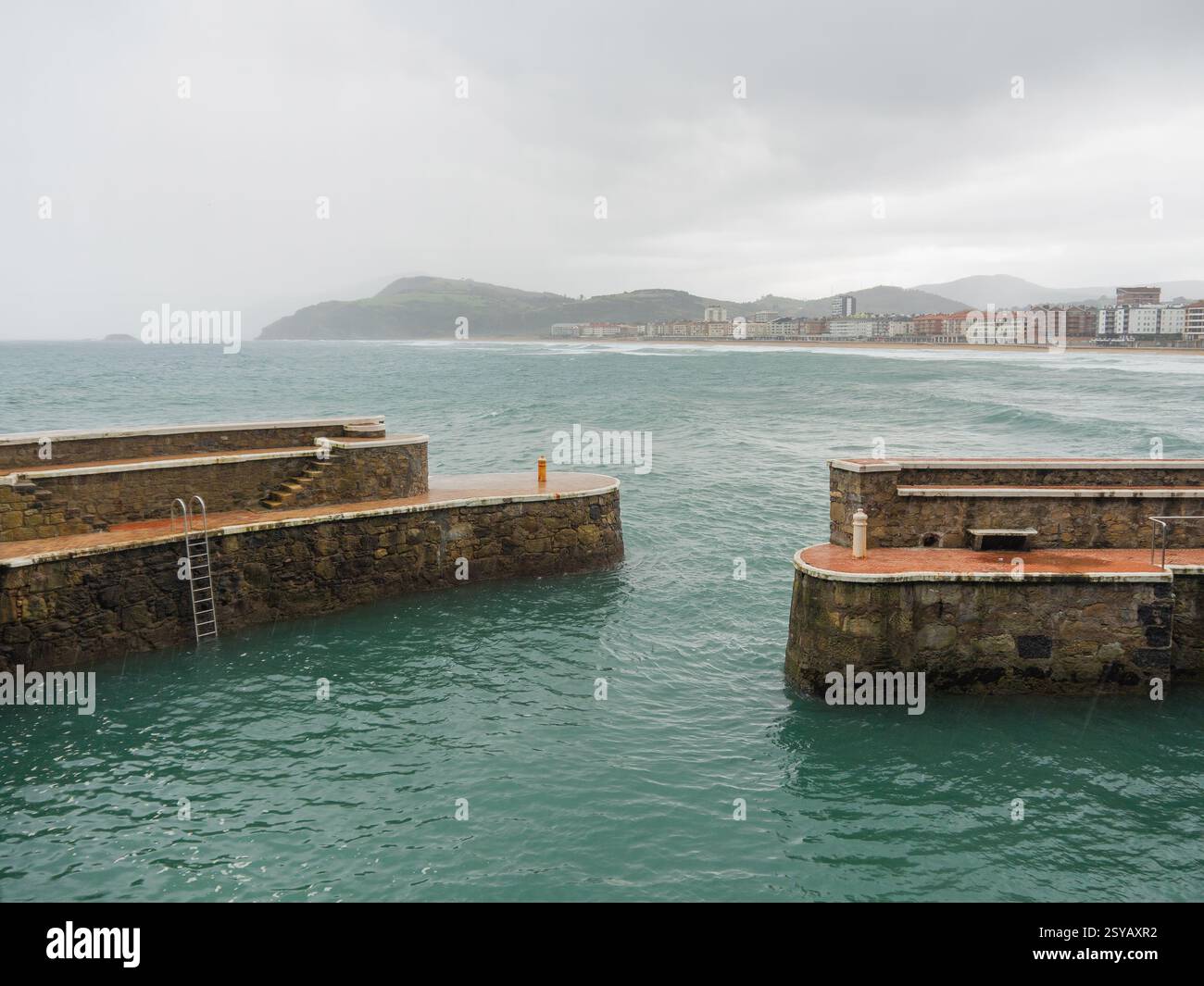 Eine beschauliche Szene an der Küste bei Regenfällen, die Steinbrüche und das turbulente Meer mit einer verschwommenen Stadtlandschaft und Bergen in der Ferne darstellt, Stockfoto