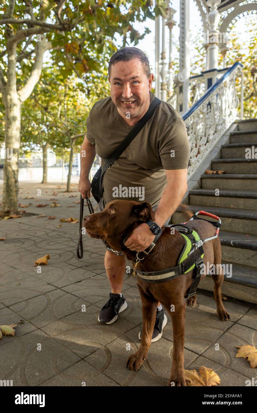 Ein lächelnder blinder Mann mit Blindenhund an der Leine genießt einen sonnigen Herbsttag in einem Park. Der Hund trägt ein Gurtzeug, und gefallene Blätter umgeben sie nahe dekorativ Stockfoto