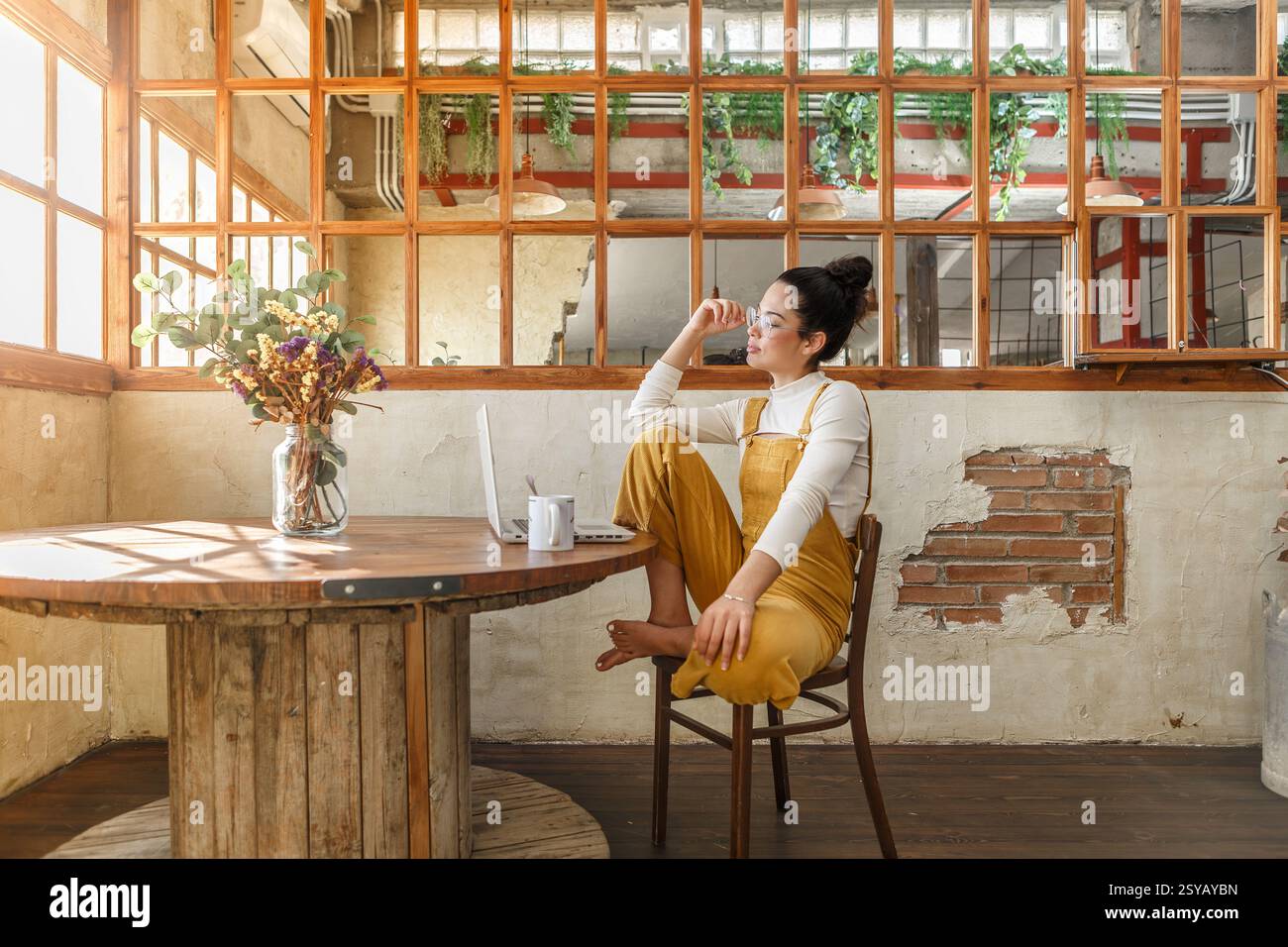 Eine junge Frau genießt eine Kaffeepause in einem sonnendurchfluteten rustikalen Loft mit einem Laptop in der Nähe, was eine entspannte Telearbeit-Umgebung veranschaulicht. Stockfoto