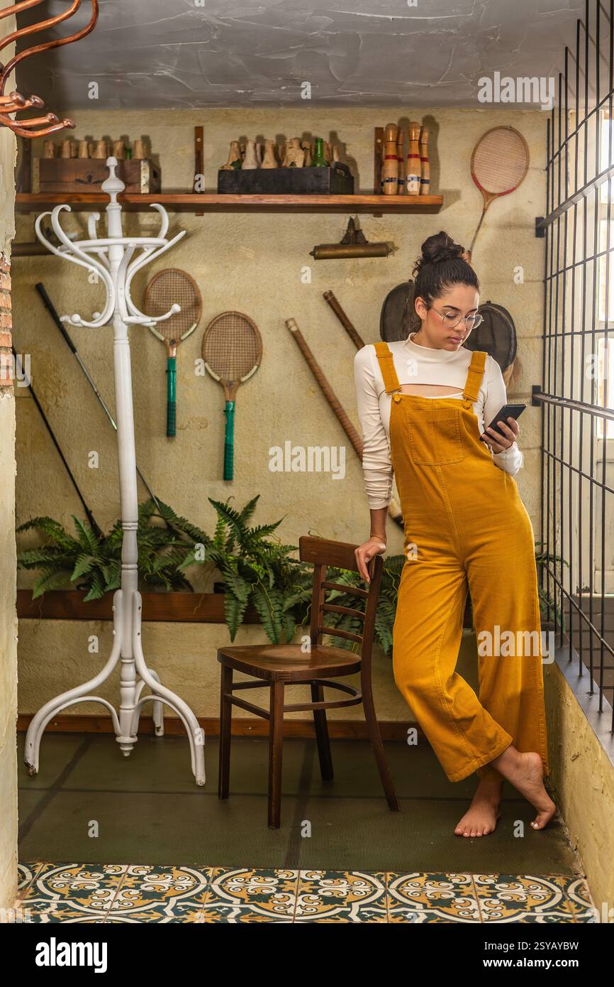 Eine junge Frau, die in modischen gelben Overalls und Brillen gekleidet ist, benutzt ein Smartphone, während sie in einem rustikalen Innenbereich mit Vintage-Sportgeräten steht Stockfoto