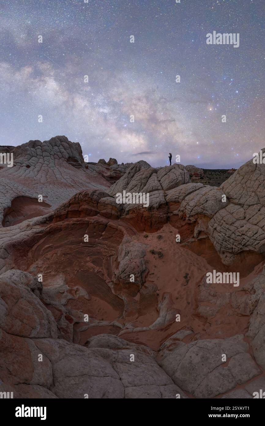 Ein weit entfernter, nicht erkennbarer Wanderer steht auf zerklüfteten Felsformationen unter einem strahlenden Sternenhimmel und fängt die wilde Schönheit von Utah, USA bei Nacht ein. Stockfoto