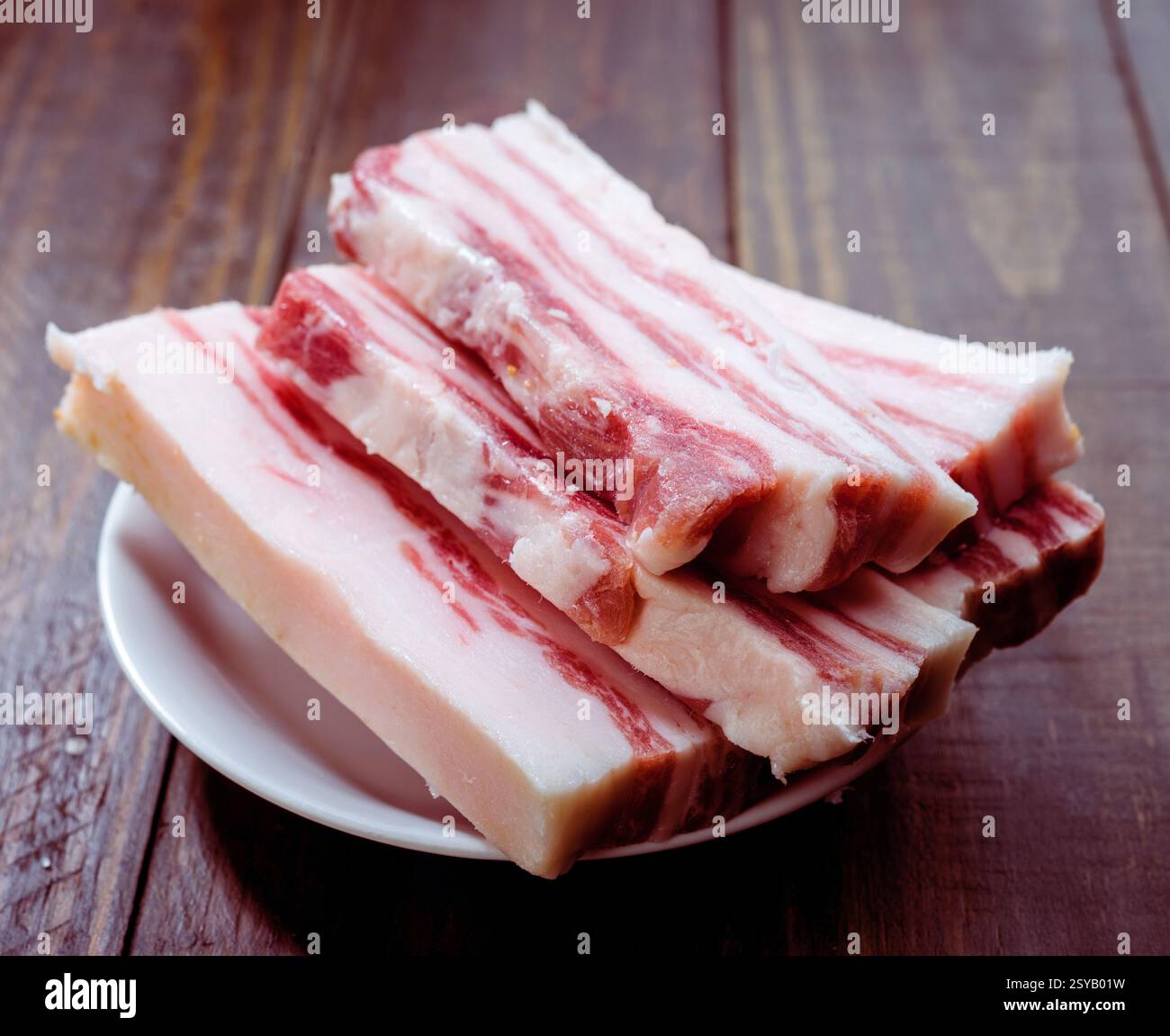 Die Scheiben von rohem Schweinebauch sind ordentlich auf einem weißen Teller auf einer rustikalen Holzoberfläche angeordnet. Das marmorierte Fett und das reichhaltige rote Fleisch betonen seine Fre Stockfoto