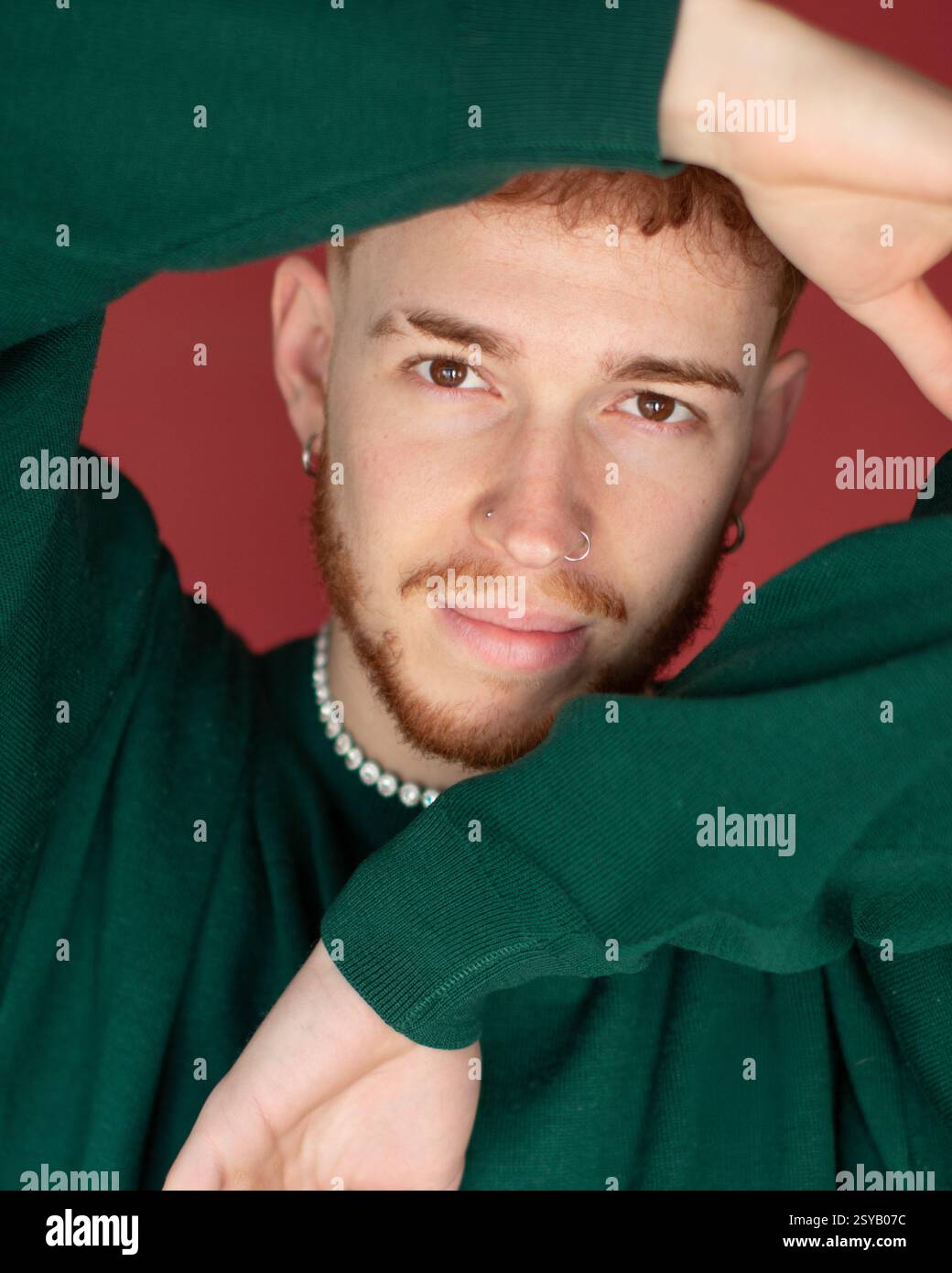 Ein Mann mit roten Haaren, die einen grünen Pullover tragen, posiert mit Armen, die sein Gesicht vor einem roten Hintergrund kreativ umrahmen. Der ausdrucksstarke Gesichtsausdruck ist hochgradig Stockfoto