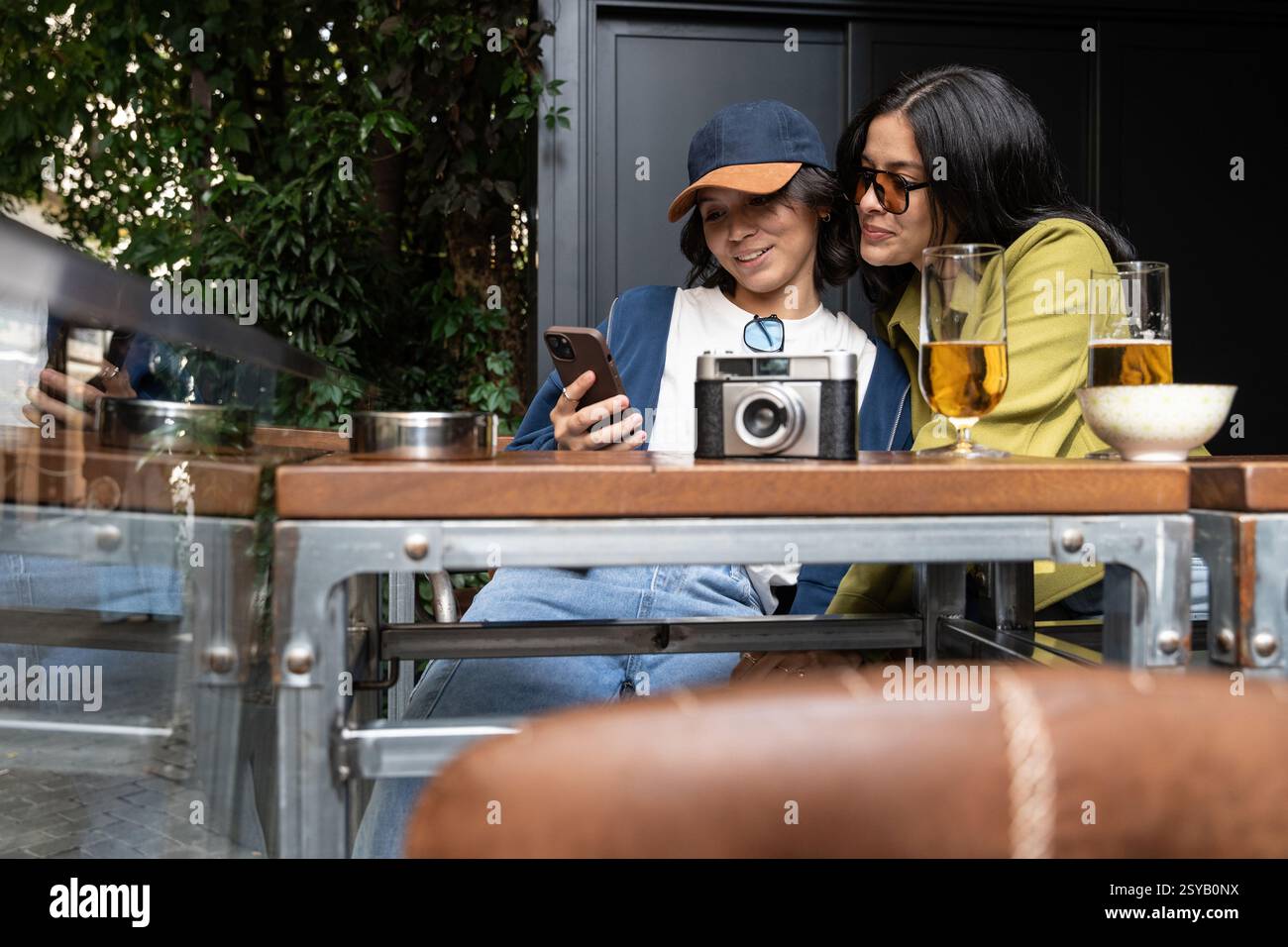 Ein Paar genießt einen entspannten Moment in einem Café und teilt ein Smartphone. Kameras und Getränke schmücken den Tisch, während üppiges Grün die gemütlichen Außenbereiche umrahmt Stockfoto