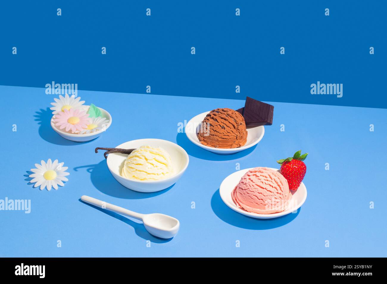 Drei verschiedene Eissorten auf einem leuchtend blauen Hintergrund, jeweils in einer weißen Schüssel mit dekorativen Elementen wie Erdbeere, Schokolade, o Stockfoto