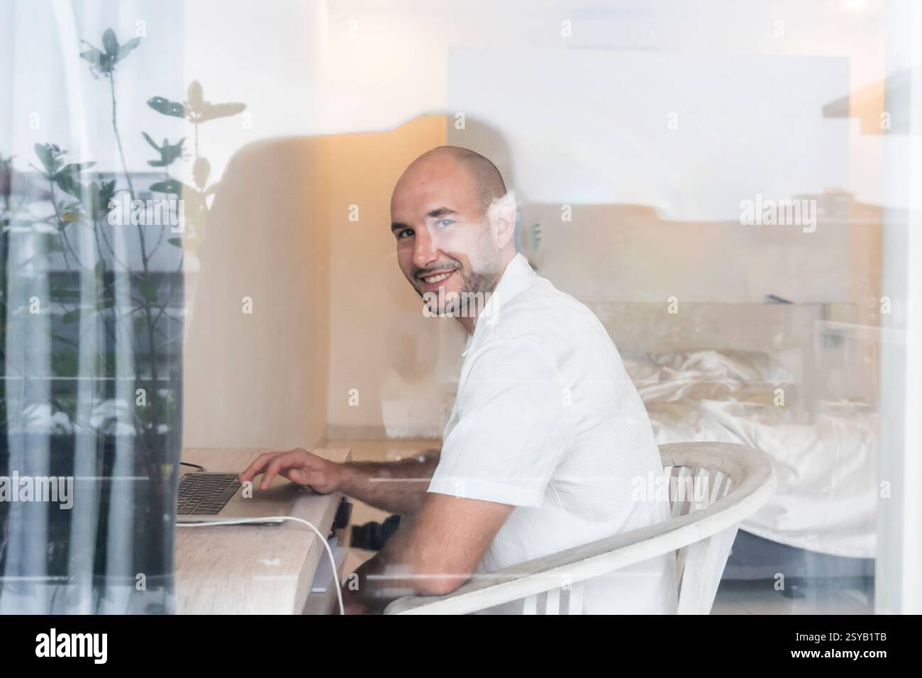 Lächelnder Mann, der in einem modernen Hotelzimmer an einem Laptop arbeitet. Natürliches Licht filtert durch das Fenster und schafft so eine ruhige Arbeitsatmosphäre, die sich positiv auf die Arbeit auswirkt Stockfoto