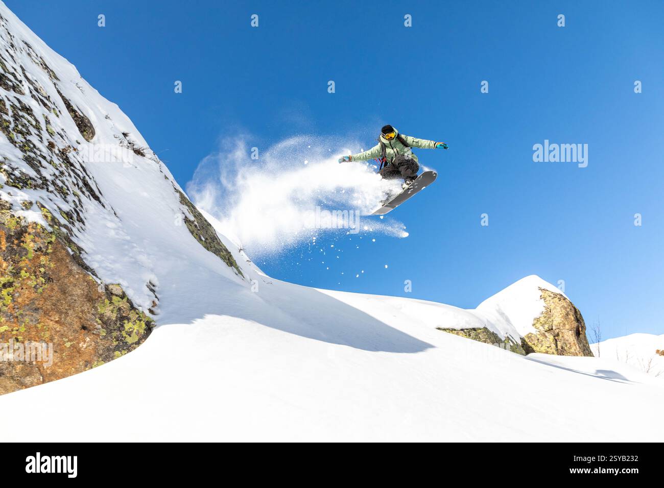 Ein Snowboarder führt einen beeindruckenden Sprung durch und schickt einen Schneespray. Die Szene spielt vor einem leuchtend blauen Himmel und schneebedeckten Bergen, Cap Stockfoto