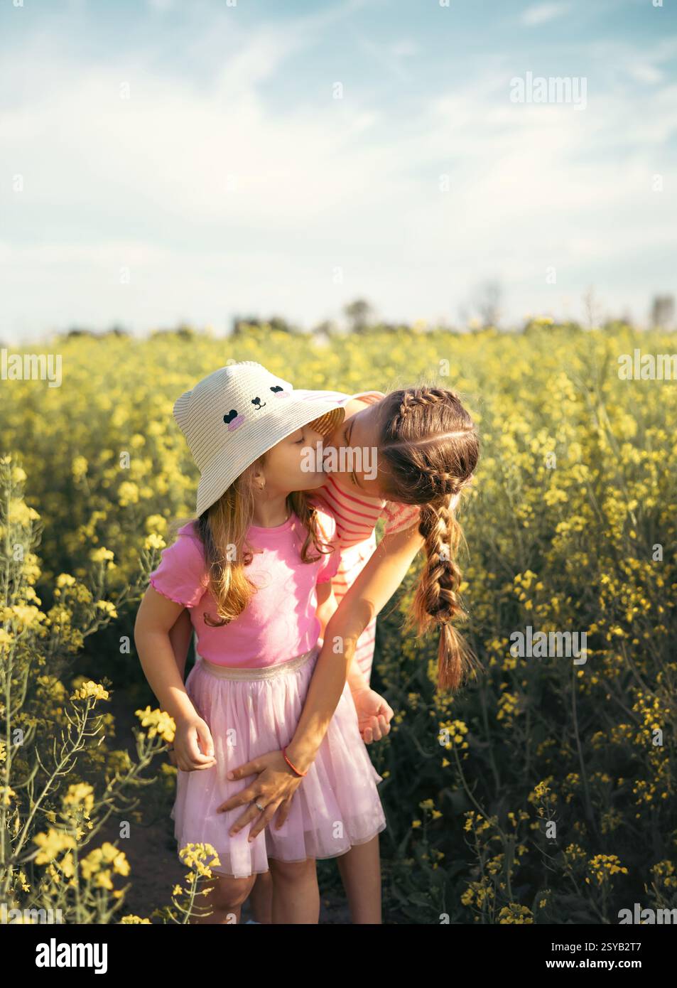 Zwei junge Schwestern teilen sich einen zarten Kuss in einem schönen gelben Blumenfeld. Ihre liebevolle Umarmung fängt die Unschuld, Freude und tiefe Bindung der Kindheit ein Stockfoto