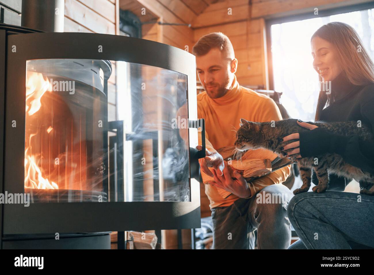 Ein Mann, der Holz in den Kamin legt. Die Frau sitzt mit einer niedlichen Katze. Stockfoto