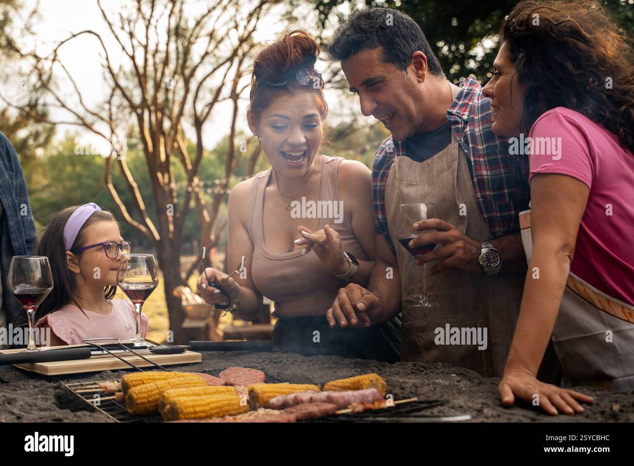 Eine fröhliche Familie und Freunde genießen ein Barbecue im Freien, teilen Essen und Lachen in einer rustikalen Umgebung mit gegrilltem Mais und Wein. Stockfoto