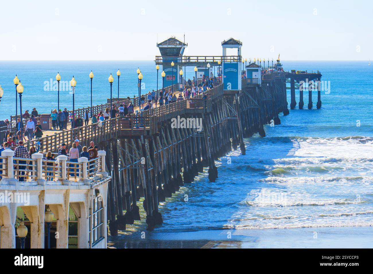 Oceanside, Kalifornien - 27. Dezember 2024: Lebhafter Oceanside Pier voller Besucher, die einen sonnigen Tag genießen. Wellen schlagen sanft gegen die Pier-Säule Stockfoto