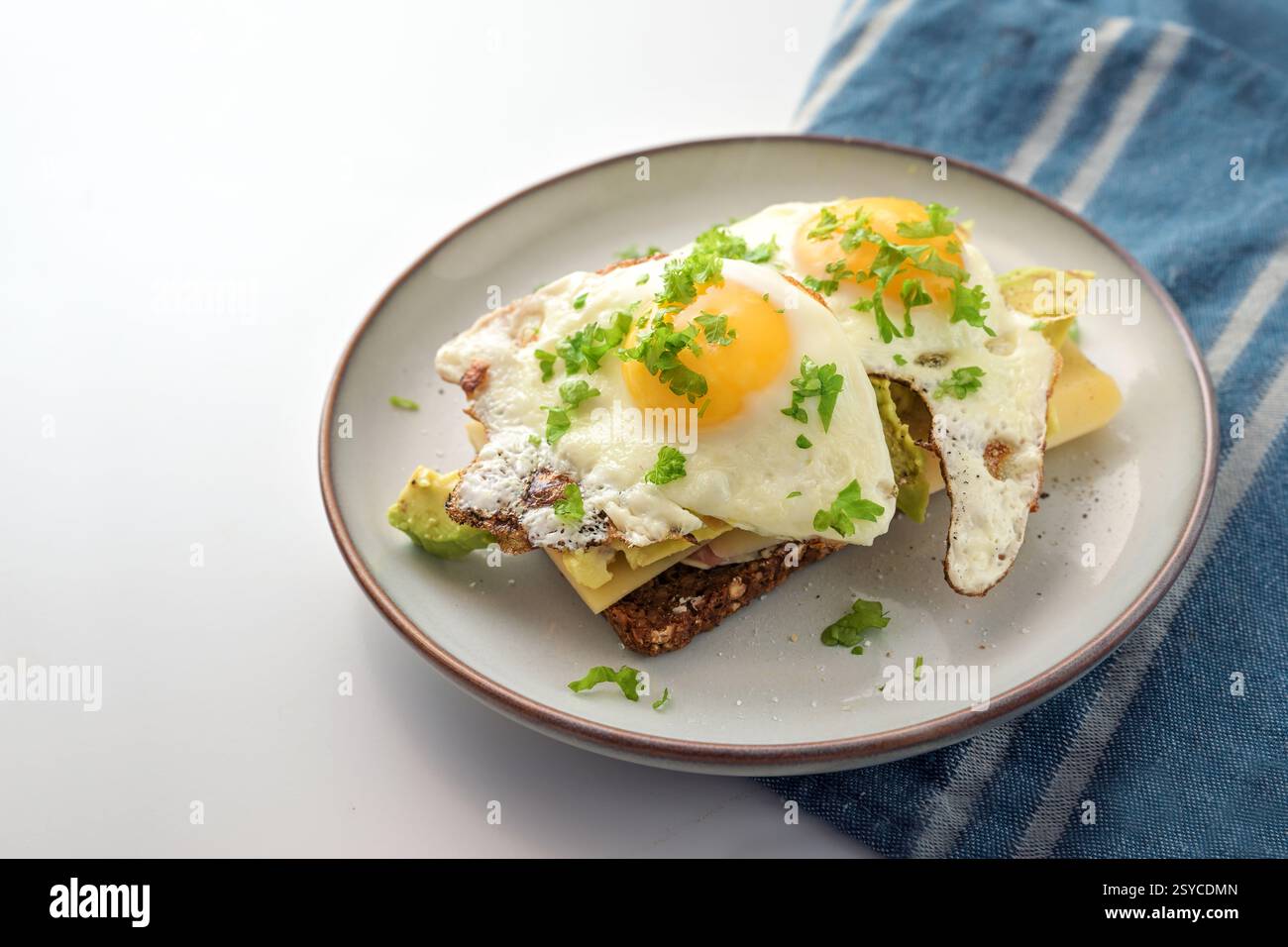 Spiegeleier auf dunklem Roggenbrot mit Käse, Avocado und Petersilie, herzhafte Mahlzeit zum Frühstück, Mittag- oder Abendessen, blaues Küchentuch, hellgraue Rückseite Stockfoto