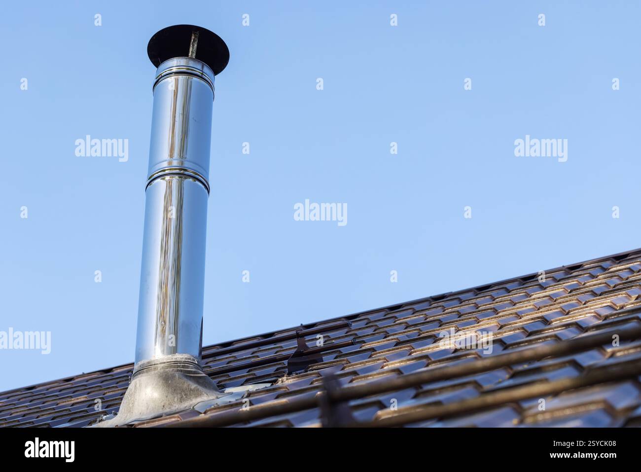 Glänzendes zylindrisches Metallschornsteinrohr mit Schutzkappe befindet sich auf dem Dach des ländlichen Hauses unter klarem blauem Himmel und erfasst Industrie- und Wohnheime Stockfoto