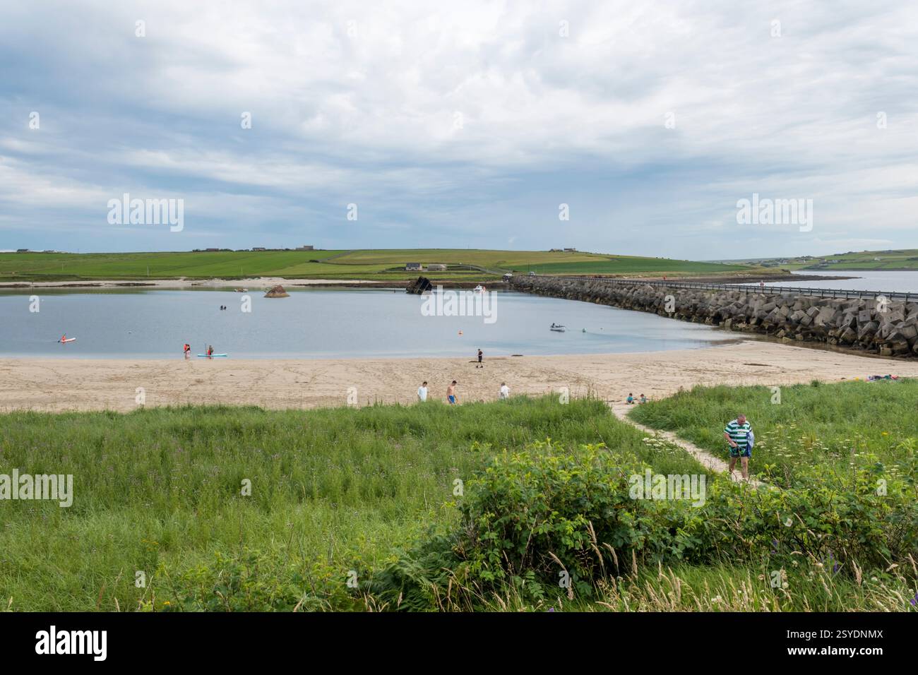 Menschen am Strand an einem sonnigen Tag neben Churchill Barrier 3 zwischen Glimps Holm und Burray, Orkney. Blockschiff im östlichen Weddell Sound im Hintergrund. Stockfoto
