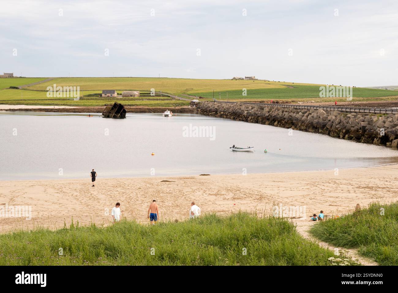 Menschen am Strand an einem sonnigen Tag neben Churchill Barrier 3 zwischen Glimps Holm und Burray, Orkney. Blockschiff im östlichen Weddell Sound im Hintergrund. Stockfoto