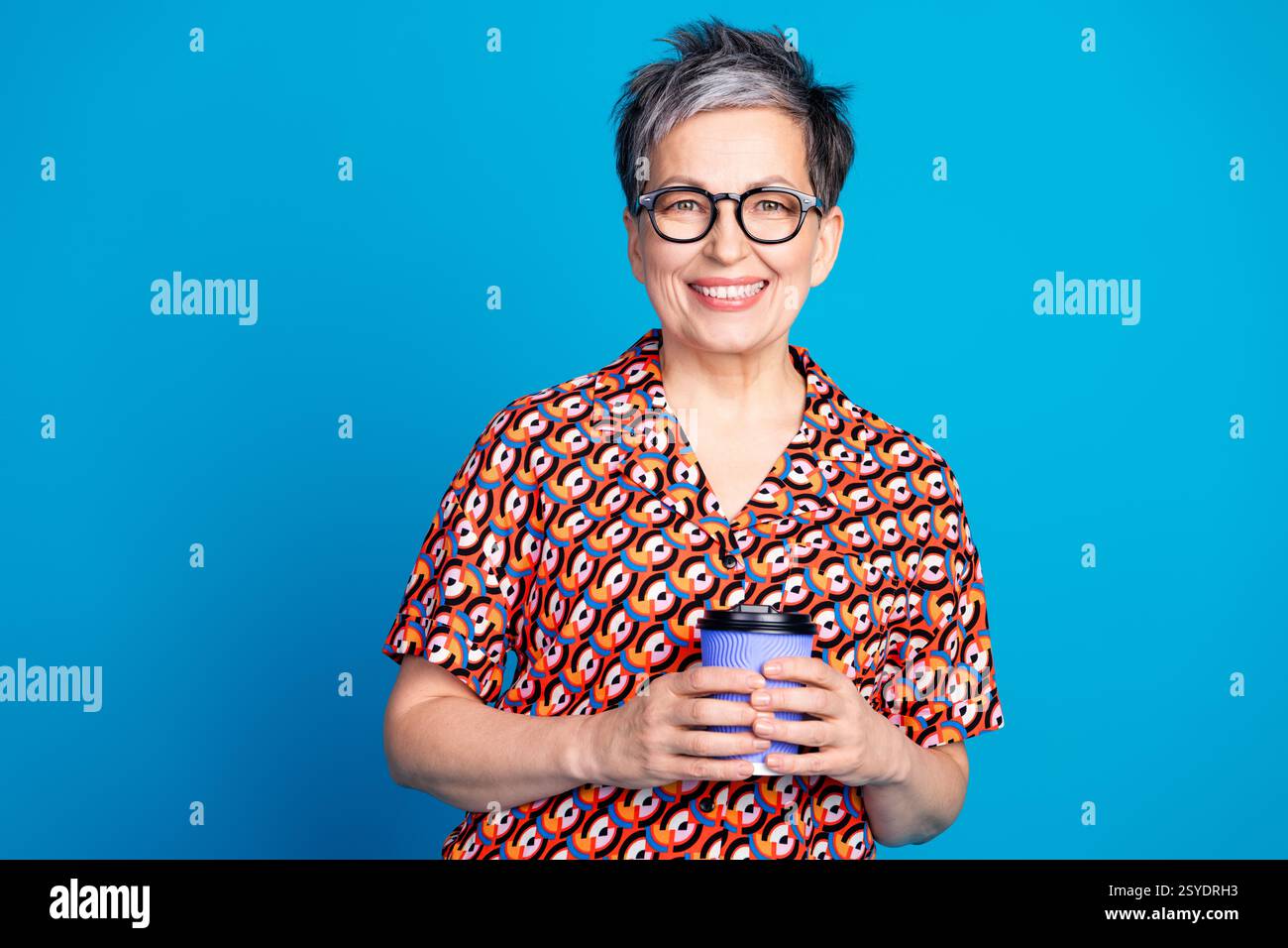 Lächelnde reife Frau in einem bunten gemusterten Shirt hält eine Kaffeetasse vor einem hellen blauen Hintergrund Stockfoto