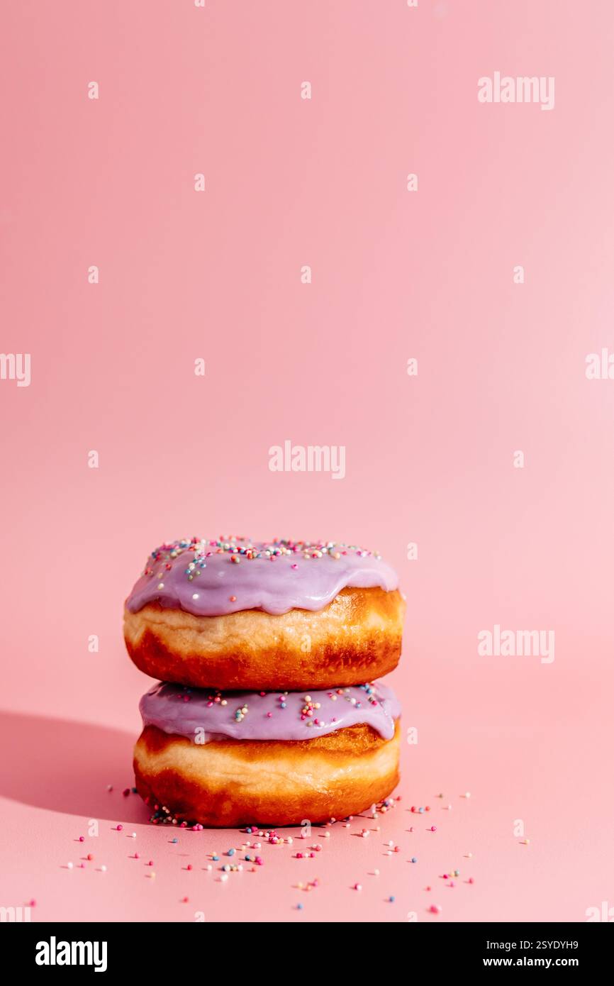 Zwei glasierte Donuts mit violettem Zuckerguss und bunten Streuseln auf einem massiven rosafarbenen Hintergrund. Stockfoto