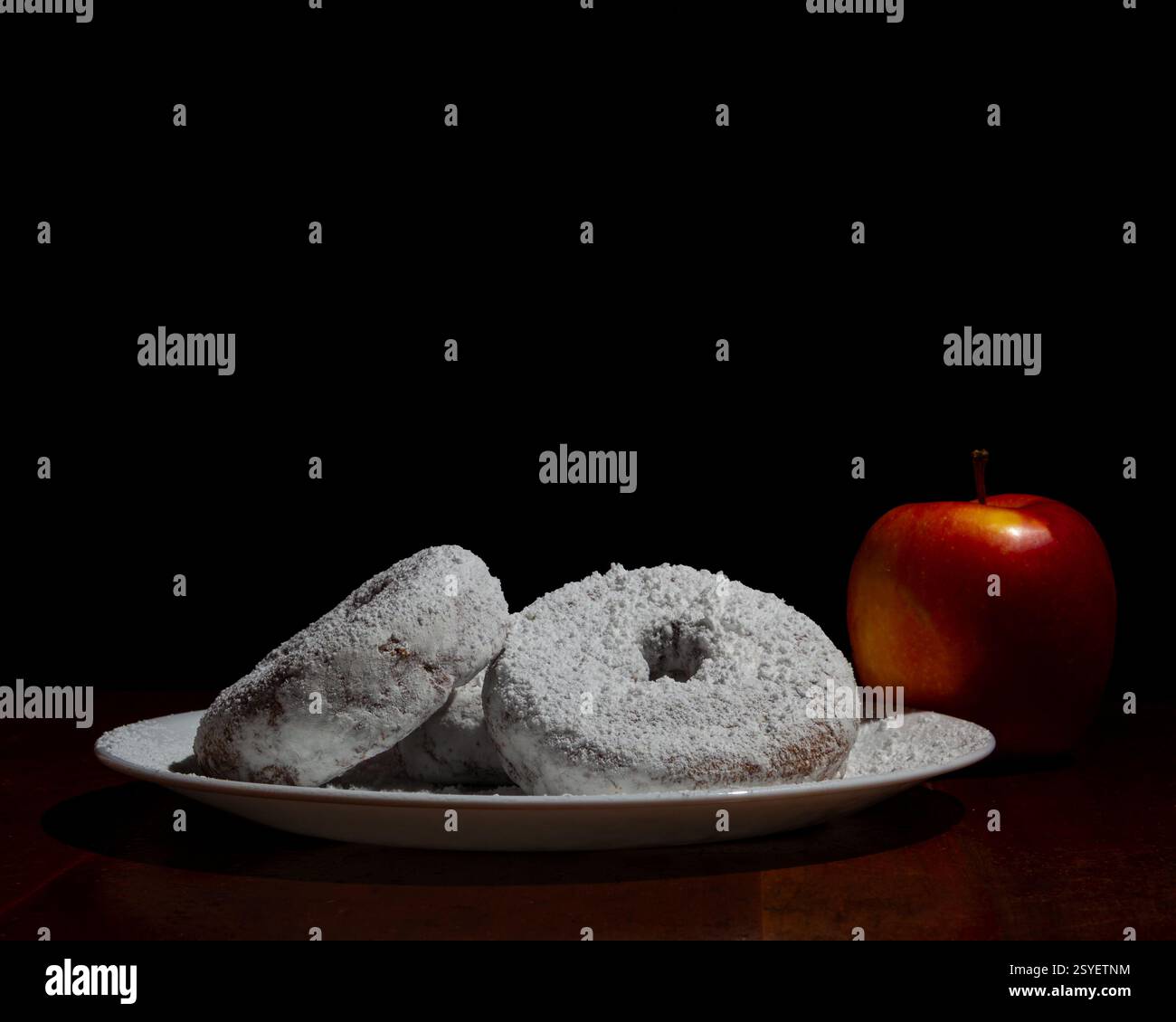 Eine Nahaufnahme von Donuts in Pulverform auf einer Keramikplatte, begleitet von einem roten Apfel, vor dunklem Hintergrund, wodurch eine stimmungsvolle Komposition entsteht. Stockfoto