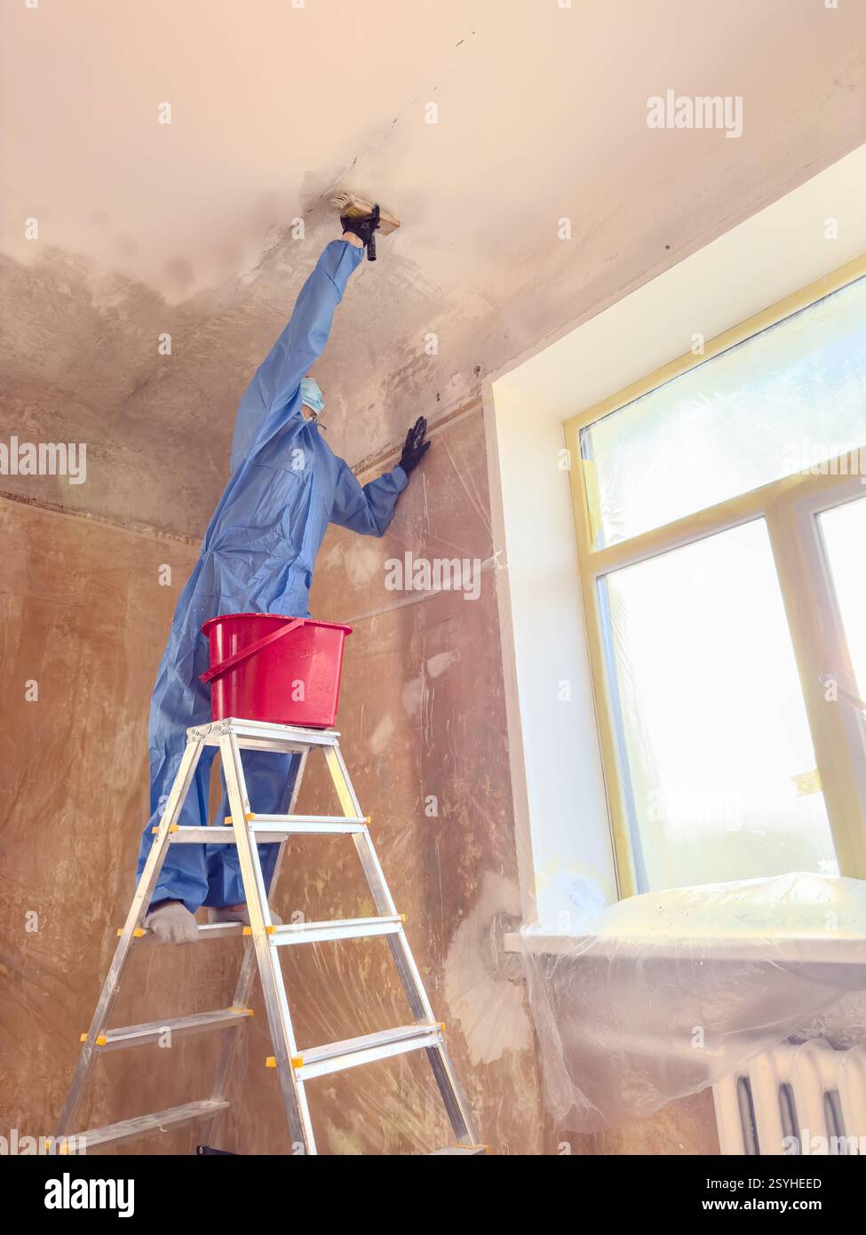 Ein Arbeiter in einem blauen Anzug wäscht gekonnt Kreide und Kalk von der Decke, während er auf einer Leiter in der Nähe eines Fensters balanciert Stockfoto Ein Arbeiter in einem blauen Anzug wäscht gekonnt Kreide und Kalk von der Decke, während er auf einer Leiter in der Nähe eines Fensters balanciert Stockfoto