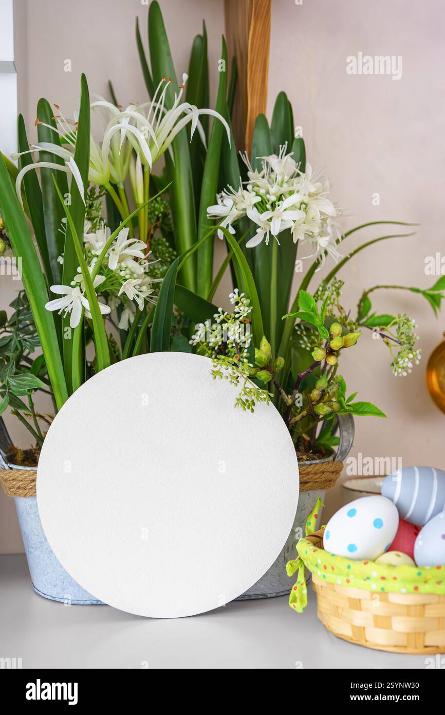 Dekorativer Topf gefüllt mit üppig grünen Pflanzen, weißen Blumen und bunten Ostereiern in Korb auf einem Regal mit leerem Kreis, leerer runder Displaykarte Stockfoto
