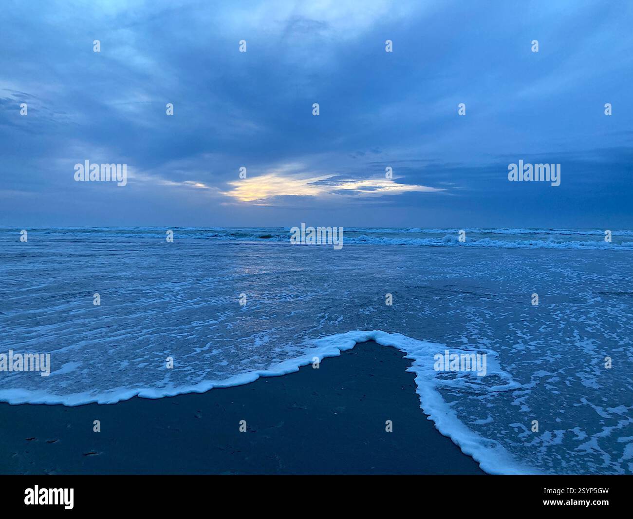 Zwei Wellen verschmelzen über nassem Sand an einem Winterstrand, während das letzte goldene Licht der Sonne durch die Wolken am fernen Horizont bricht. - Smartphone-aufgenommenes Stockfoto
