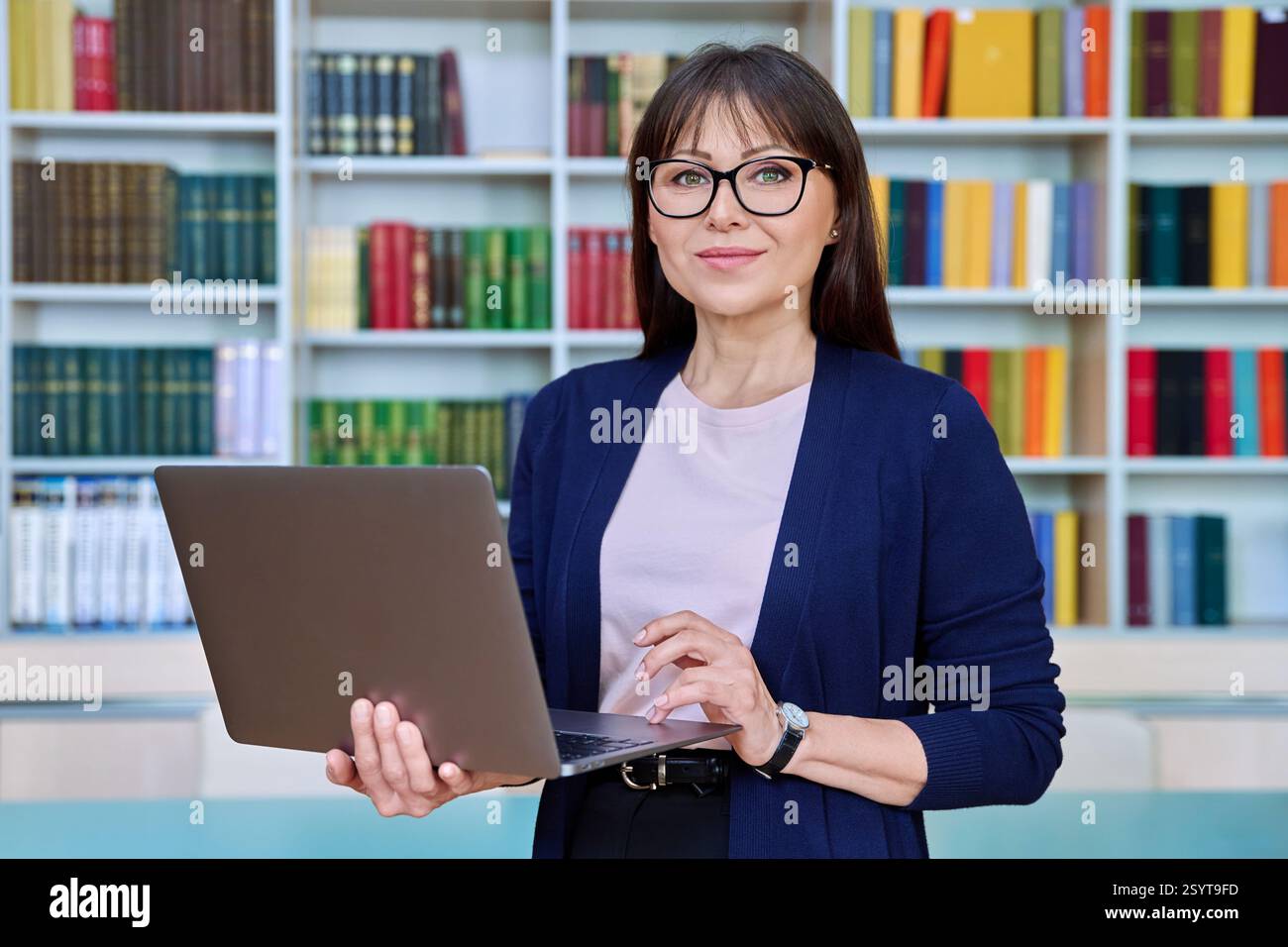 Porträt einer lächelnden Lehrerin mittleren Alters mit Laptop in der Bibliothek Stockfoto