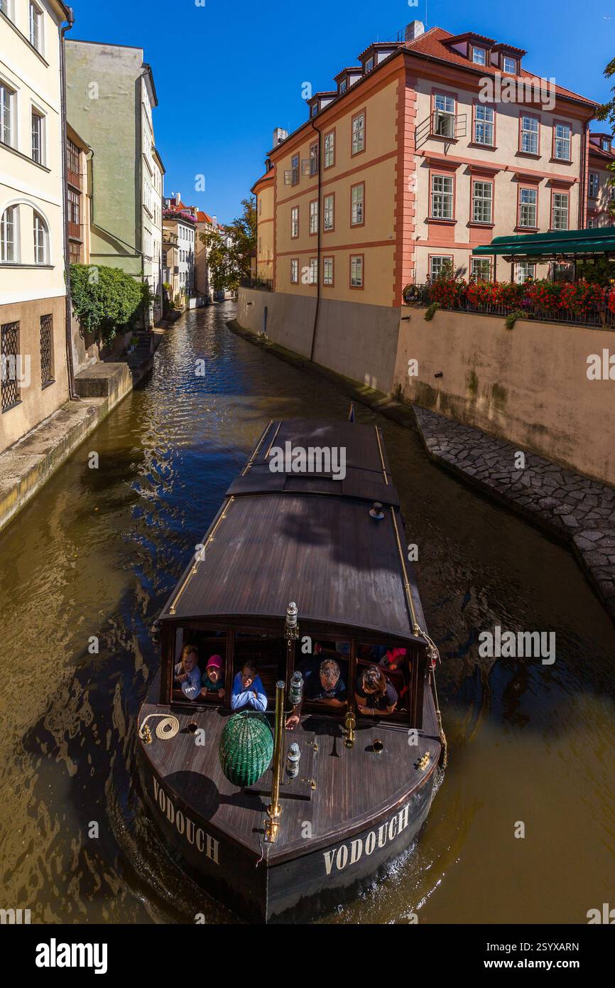 Ein schmaler Kanal, gesäumt von bunten Gebäuden in Prag, mit einem Boot, das Passagiere durch das ruhige Wasser befördert. Stockfoto
