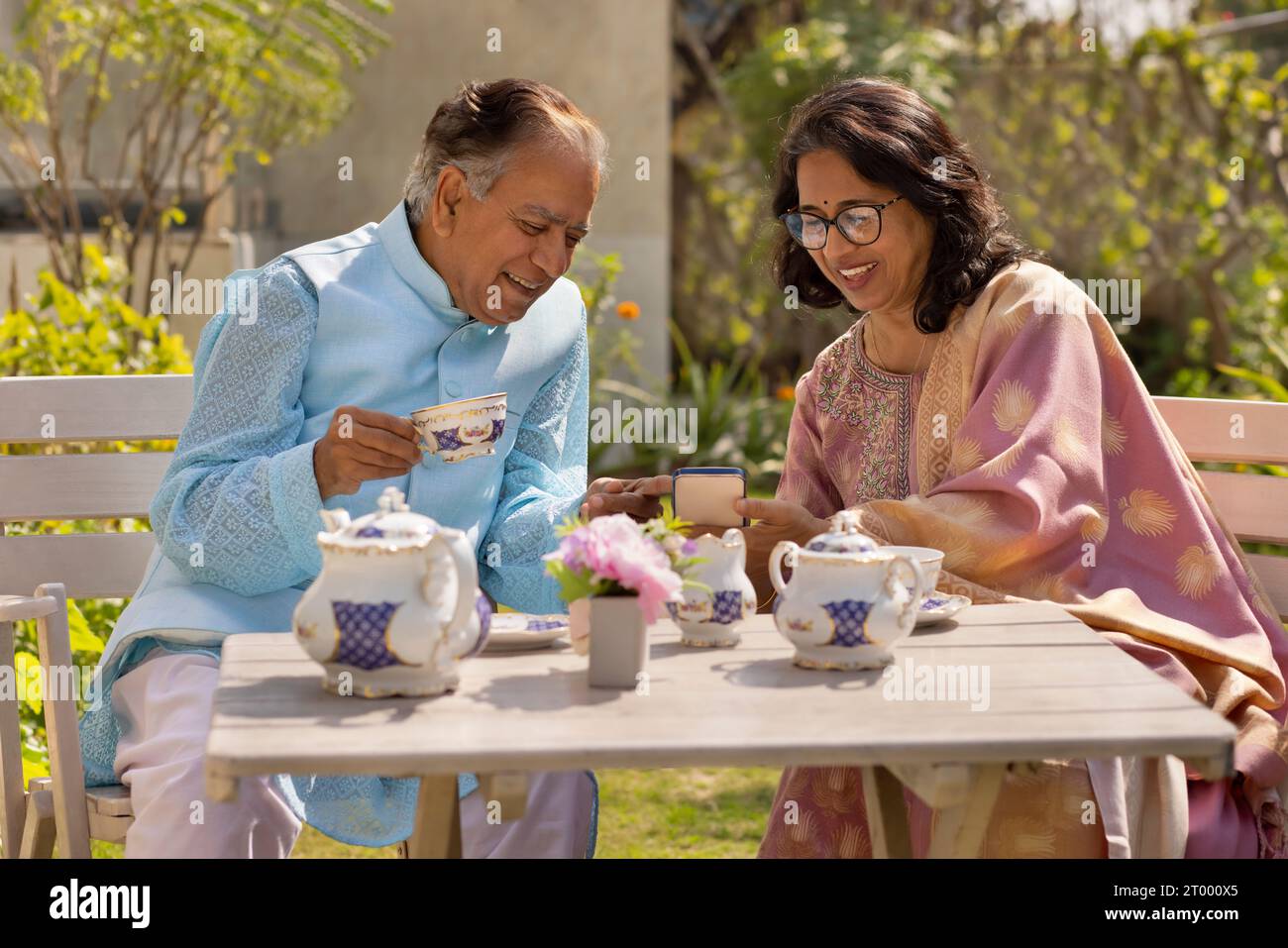 Ein älteres Ehepaar benutzt das Handy, während er im Garten Tee trinkt Stockfoto
