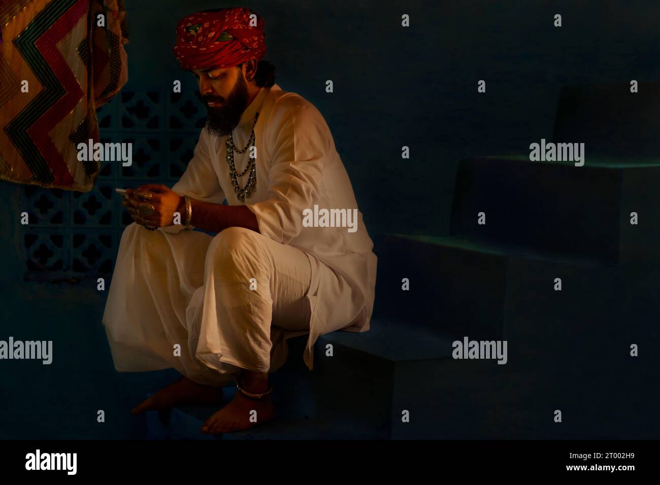 Porträt eines Rajasthani-Mannes, der das Handy benutzt, während er zu Hause auf der Treppe sitzt Stockfoto