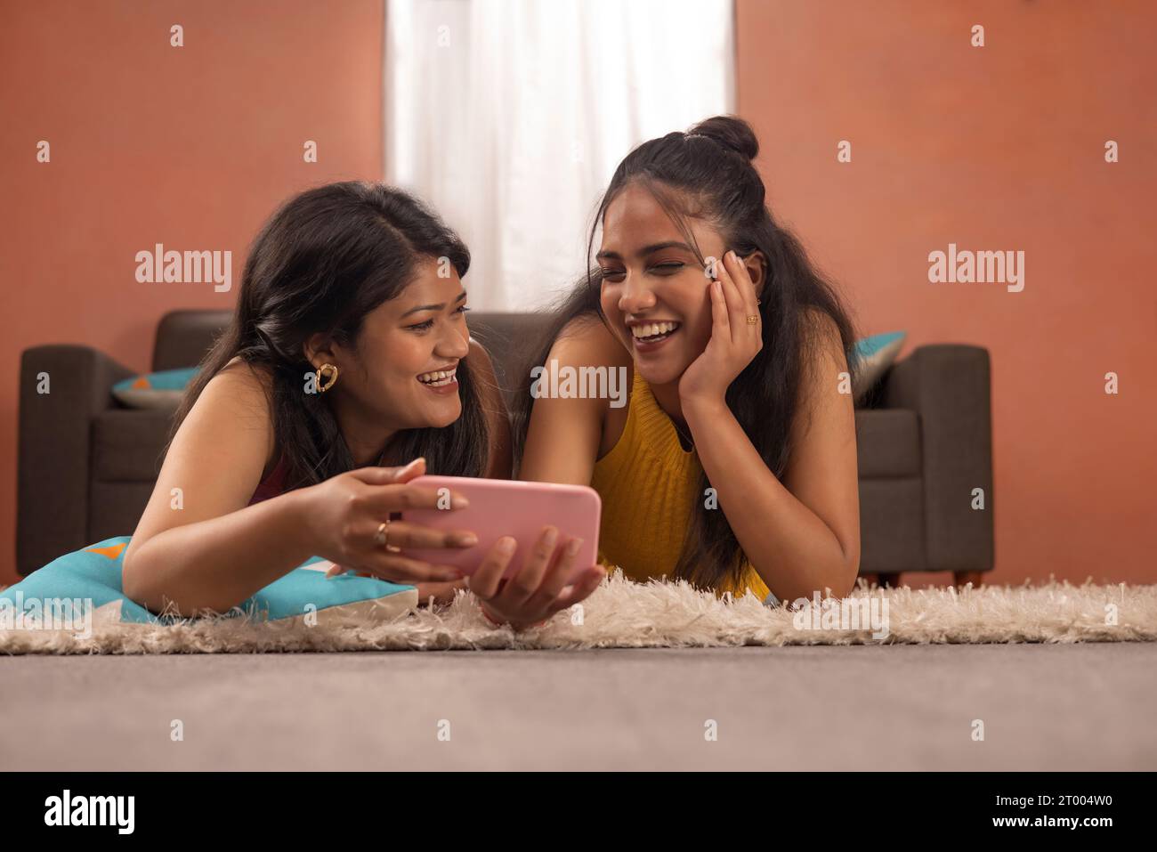 Zwei lächelnde Frauen sehen gemeinsam Videos auf dem Mobiltelefon, während sie im Wohnzimmer auf dem Boden liegen Stockfoto