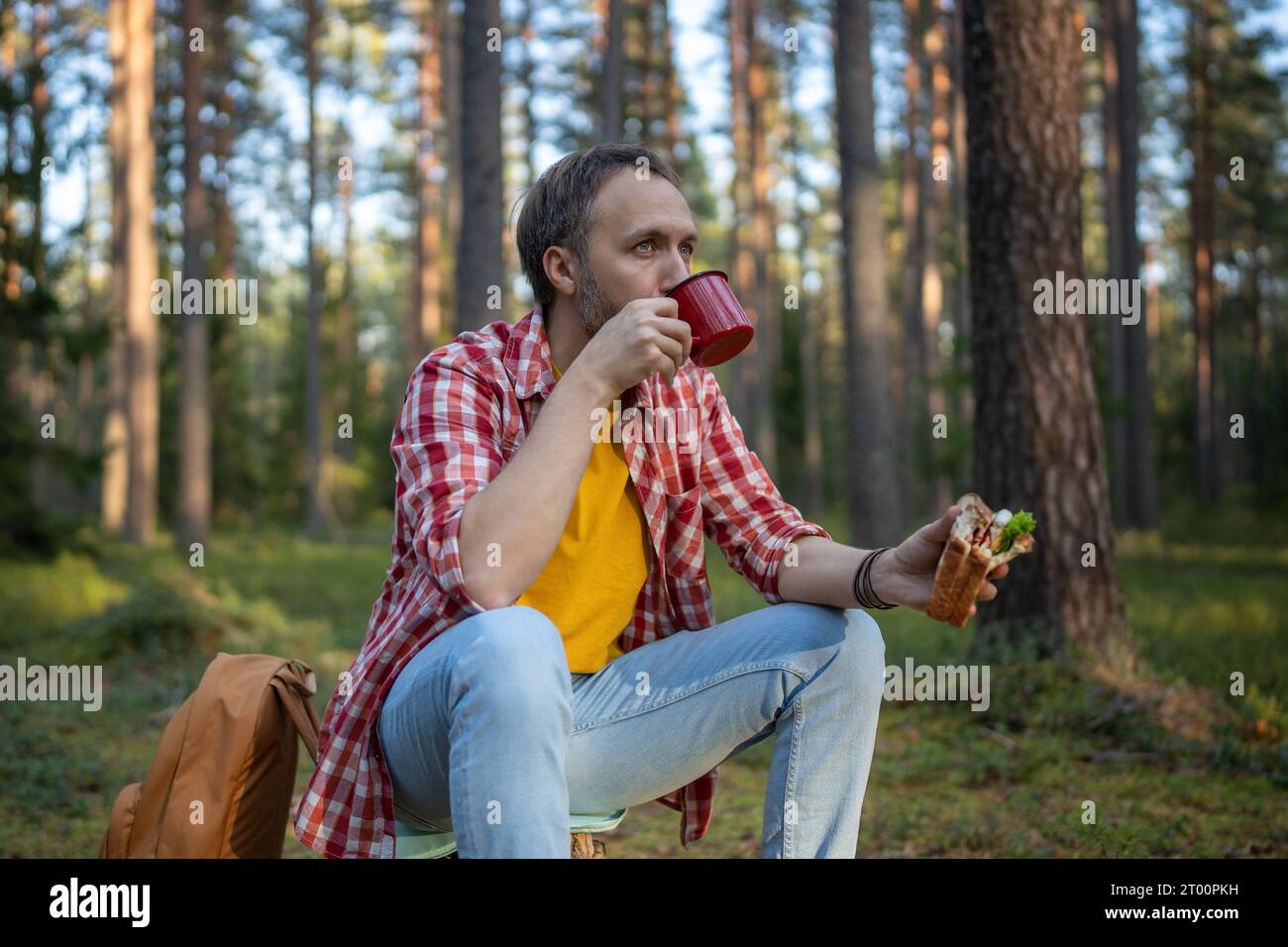 Mann Wanderer trinkt Tee aus der Tasse, isst Sandwich während der Wanderung im Wald und hält für eine Pause an Stockfoto