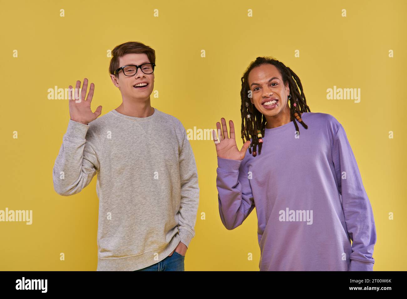 Zwei fröhliche Freunde in lila und weißen Sweatshirts, die vor der Kamera winken und lächeln, Vielfalt Stockfoto