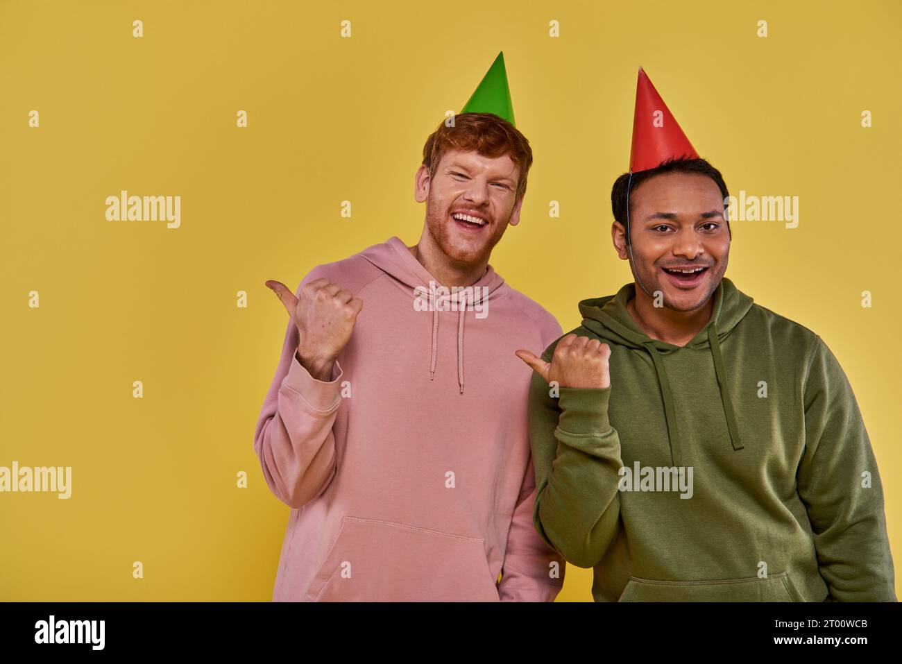 Zwei junge Freunde in lässiger Kleidung und Geburtstagshüten lächeln in die Kamera und zeigen die Daumen zur Seite Stockfoto