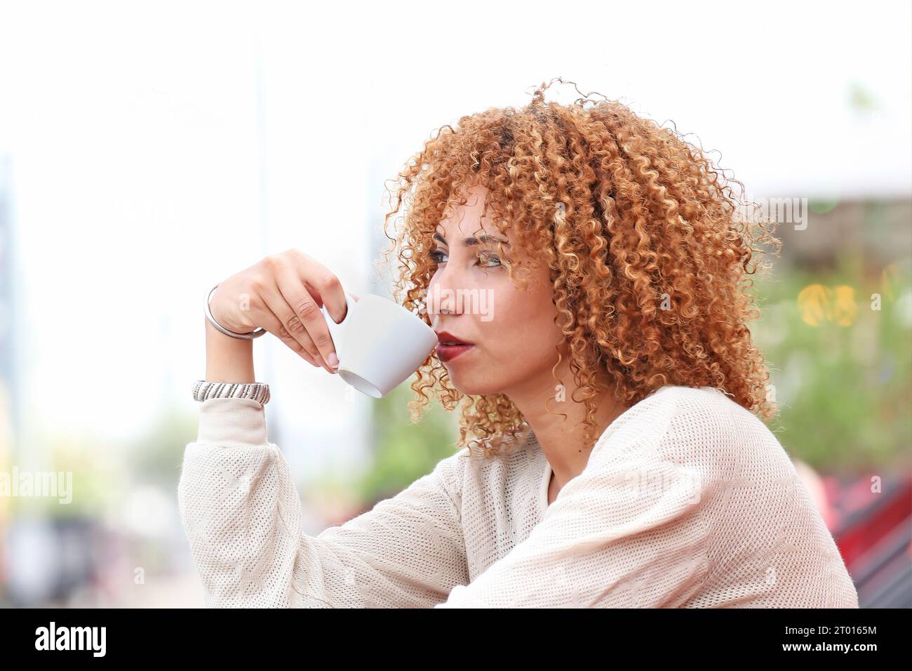 Frau mit lockigen Haaren in weißem Pullover, die draußen Kaffee trinkt und sich amüsiert Stockfoto