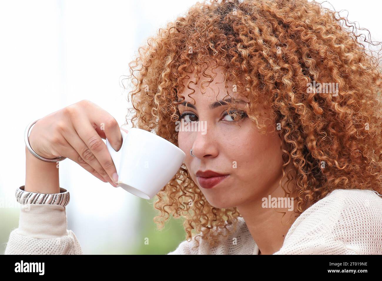 Frau mit lockigen Haaren in weißem Pullover, die draußen Kaffee trinkt und sich amüsiert Stockfoto