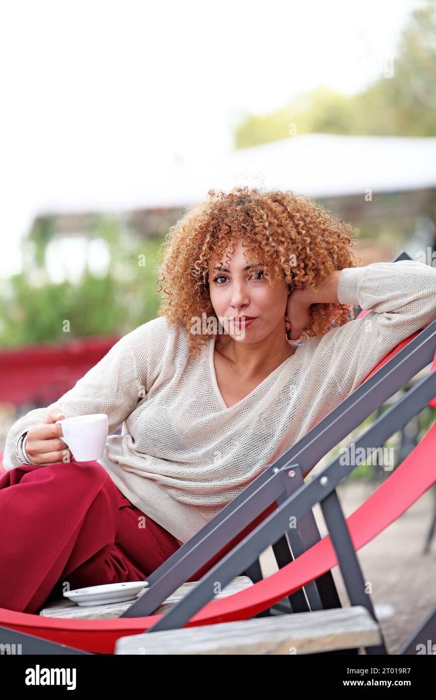 Frau mit lockigen Haaren in weißem Pullover, die draußen Kaffee trinkt und sich amüsiert Stockfoto