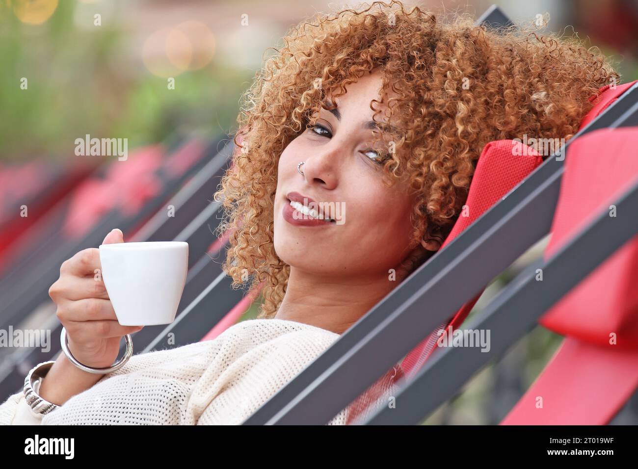 Frau mit lockigen Haaren in weißem Pullover, die draußen Kaffee trinkt und sich amüsiert Stockfoto