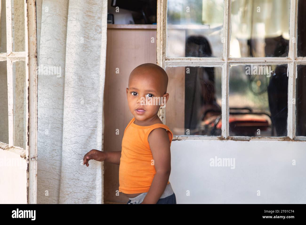 afrikanisches Kind vor dem Haus im Township, in einer orangefarbenen Weste, vor der Tür stehend Stockfoto