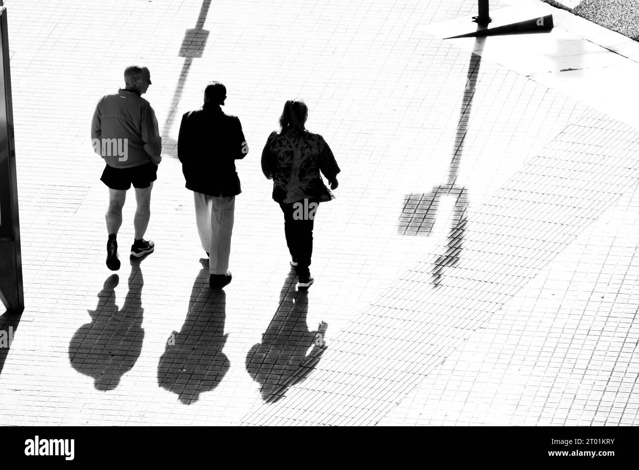 Drei Leute laufen die Straße runter. Das starke Licht hebt ihre Silhouetten und ihre Schatten auf dem Boden hervor. Stockfoto