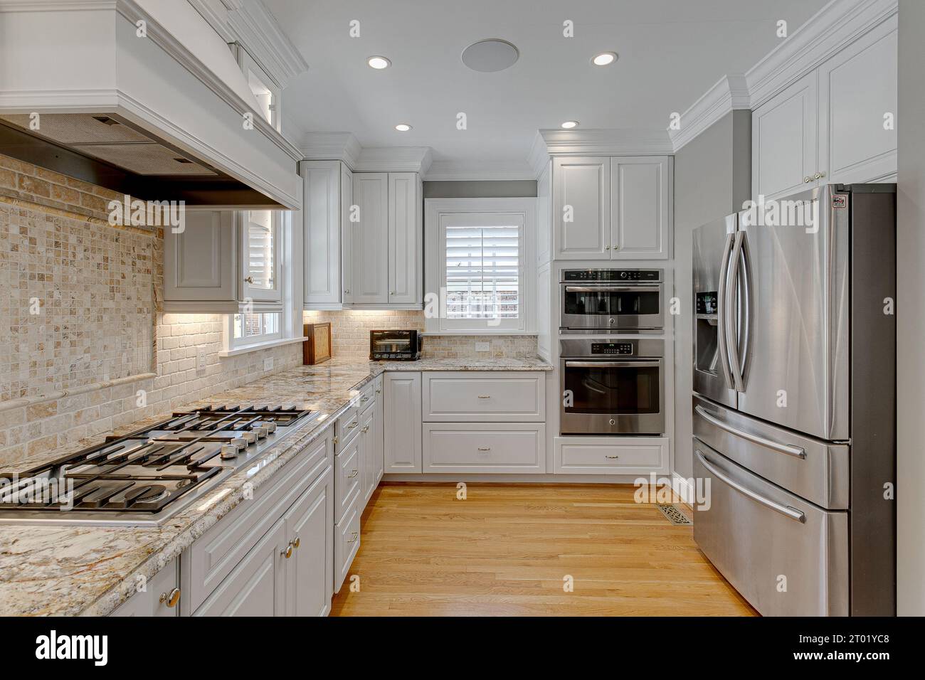 Moderne, saubere und luxuriöse weiße Küche mit Herdplatte mit Backsplash und Edelstahlgeräten mit Hartholzböden und Rollläden Stockfoto