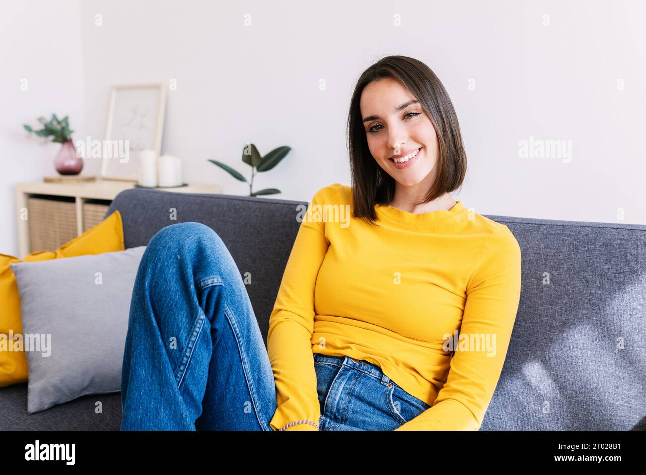 Schöne junge Erwachsene kaukasische Frau lächelt in die Kamera und sitzt zu Hause auf dem Sofa Stockfoto
