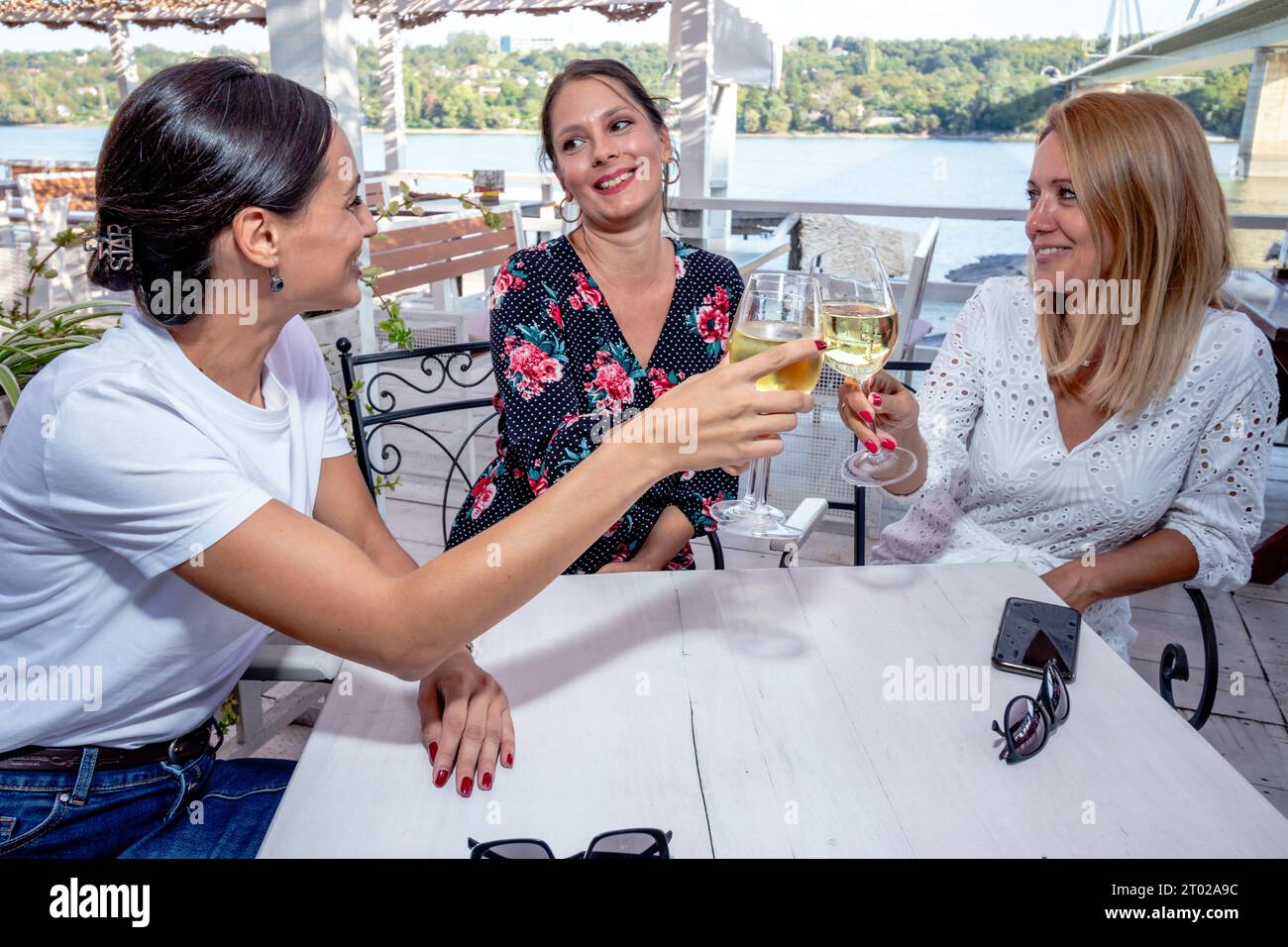 Drei fröhliche Freundinnen sitzen an einem Tisch mit einem Glas Wein, toasten, plaudern und lachen auf einer Restaurantterrasse am Fluss. Stockfoto