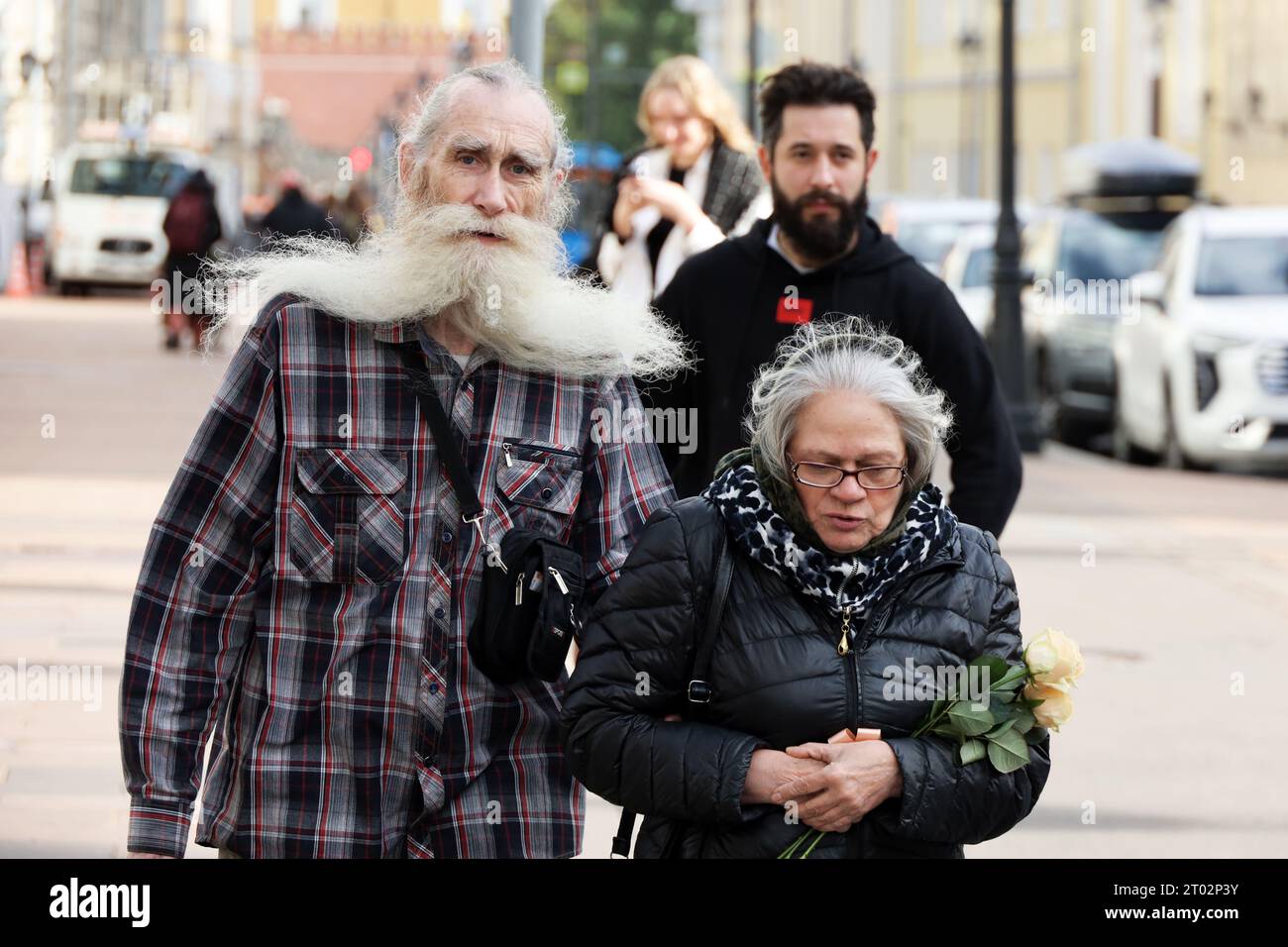 Ein älteres Paar, das auf der Stadtstraße läuft und Hände hält. Mann mit langem weißem Bart und Frau mit Blumenstrauß Stockfoto