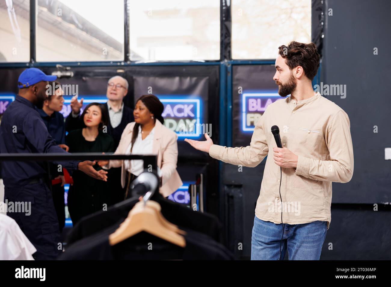 Man TV-Reporter, der am Eingang des Einkaufszentrums steht und Nachrichten über Black Friday präsentiert. Der Sicherheitsdienst des Bekleidungsgeschäfts öffnet die Tür für eifrige Leute, die auf Rabatte und saisonale Verkäufe warten Stockfoto