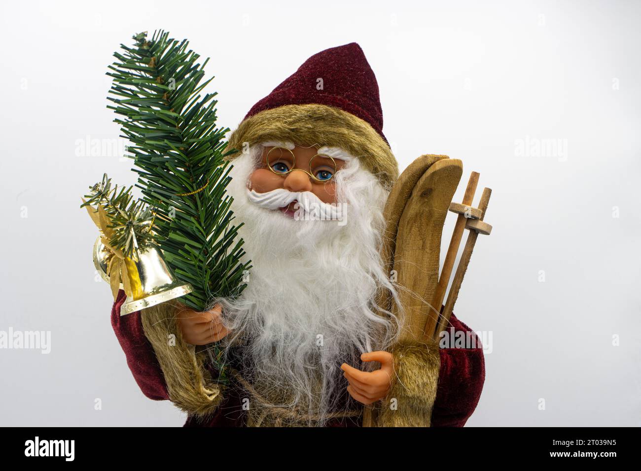 Das Weihnachtsmann-Spielzeug hält Holzskier in der einen Hand und einen Tannenzweig mit einer Glocke in der anderen auf weißem Hintergrund Stockfoto